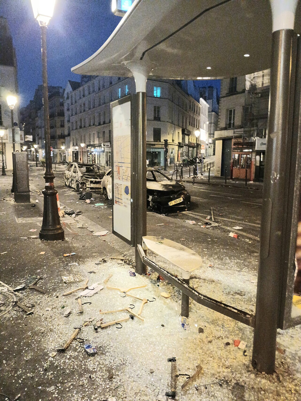 Burned cars behind a bus shelter with shattered glass and debris on the pavement.